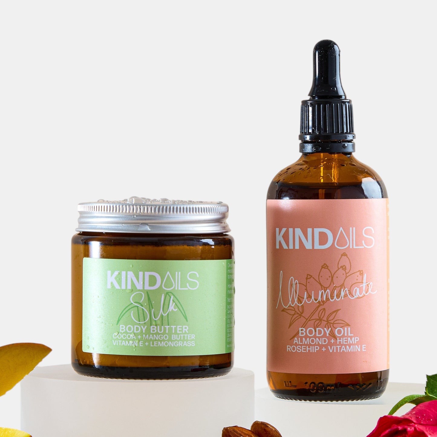 Two skincare products from kind oils  on a white background with ingredients like almonds and a rose.
