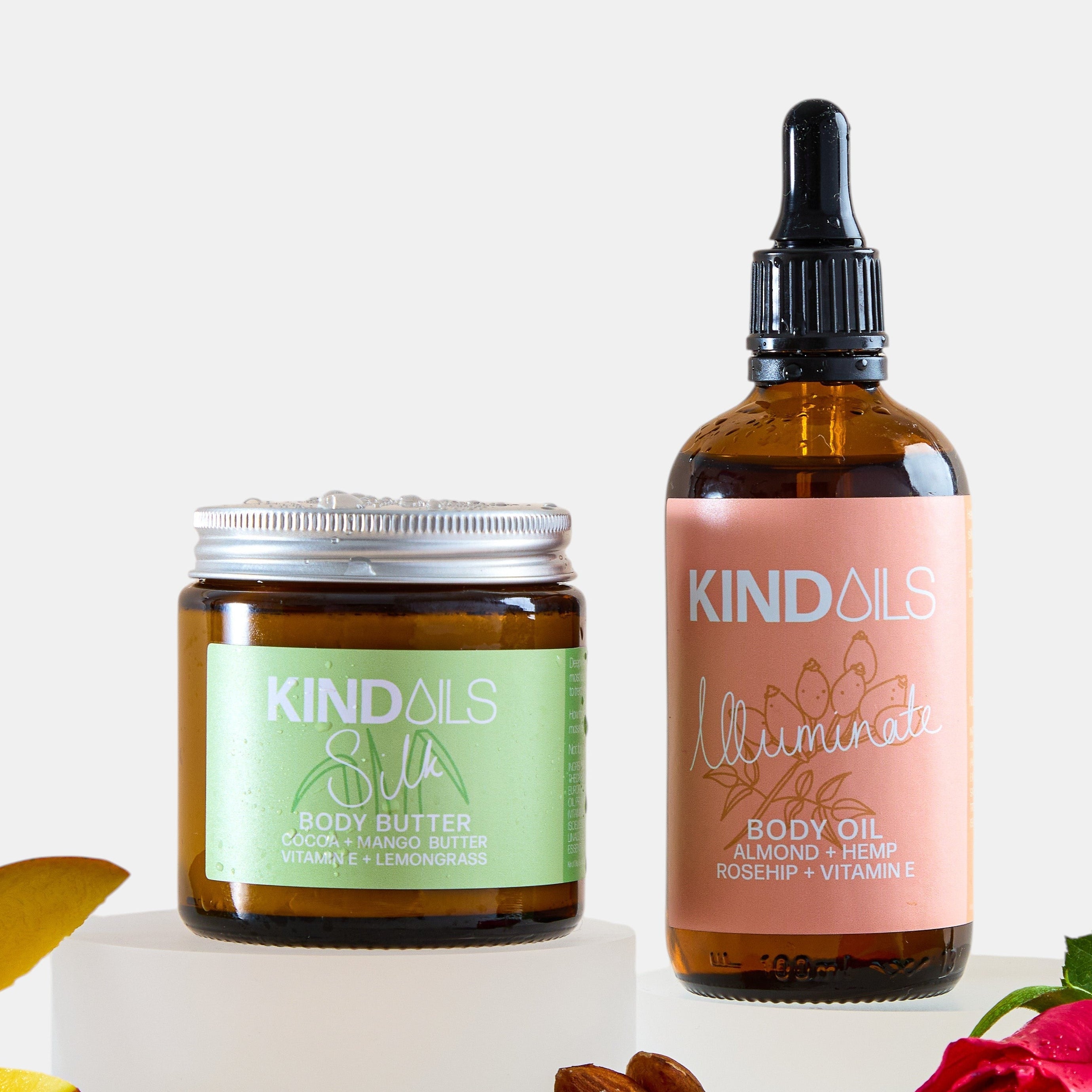 Two skincare products from kind oils  on a white background with ingredients like almonds and a rose.
